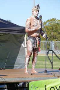 Aboriginal Man in traditional outfit for NAIDOC Week on. stage giving a speech.